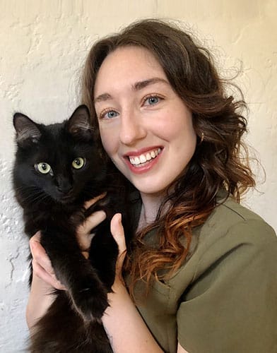 A woman with wavy brown hair smiles at the camera while holding a fluffy black cat with green eyes. The background is a pale, textured wall.