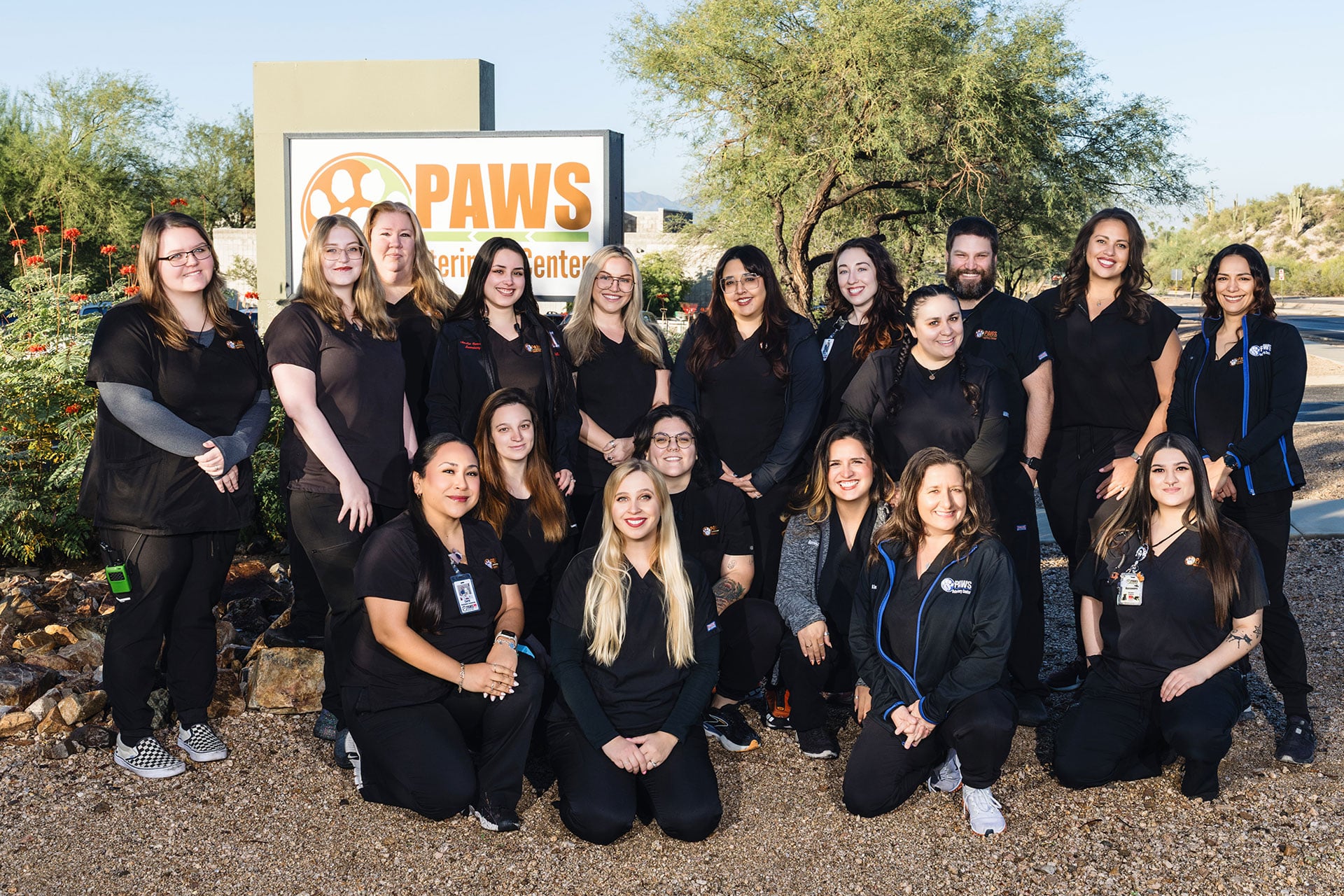 A group of 17 people dressed in black scrubs pose and smile in front of a PAWS Veterinary Center sign outdoors, with trees and desert plants in the background.