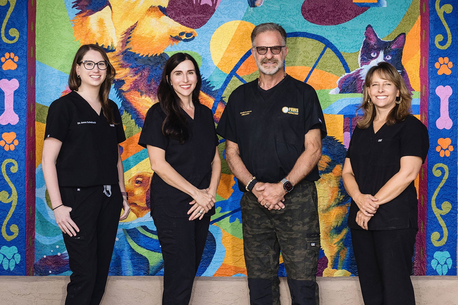Four veterinarians in black uniforms stand in front of a colorful mural featuring animals and abstract designs. They are smiling and standing close together. The mural shows a dog, cat, and various vibrant patterns.