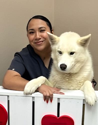 A woman in dark scrubs smiles while standing beside a white dog with pointy ears, both leaning over a white fence decorated with red hearts.