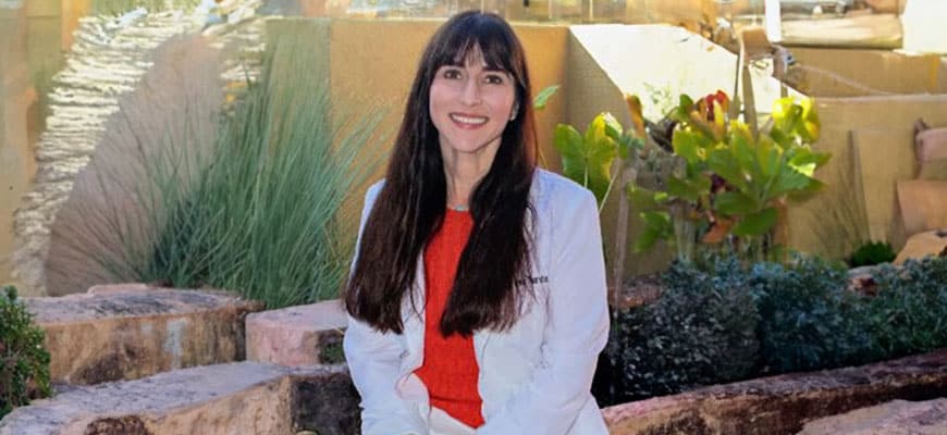 A person with long dark hair sits outdoors on a stone bench, wearing a white coat over a red top. The veterinarian is smiling, with greenery and a textured stone wall in the background.