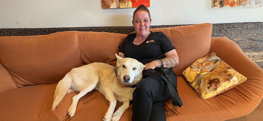 A woman with tattoos sits on an orange couch next to a light-colored dog, embodying the relaxed confidence of a veterinarian after a long day. She's wearing black clothes and smiling at the camera while the dog lies comfortably beside her. Colorful paintings adorn the wall behind them.