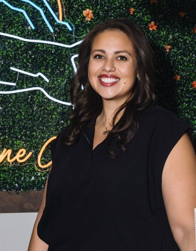 Diana, a smiling woman with long dark hair in a black top, stands in front of a green plant wall.