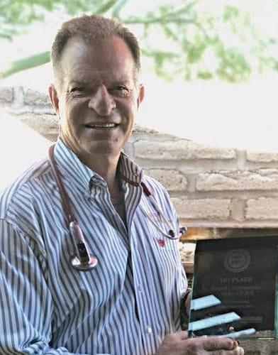 A man wearing a striped shirt and stethoscope, clearly a skilled veterinarian, smiles while holding an award. He stands proudly in front of a light brick wall with greenery in the background, showcasing his dedication to animal care.