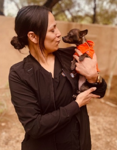A person wearing a black jacket holds a small dog dressed in an orange outfit, looking like they just came from the vet. They are outside, standing close to a wall, and appear to be playfully interacting as the person makes a playful face at the dog.