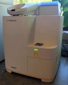A white medical or laboratory device with a digital screen and various buttons, possibly used for analysis or testing by a veterinarian, is positioned on a countertop. The surrounding environment includes a window with partial sunlight.