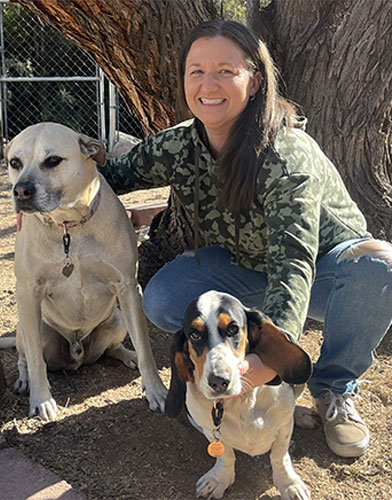 A person in a camo hoodie, who might just be your friendly neighborhood vet, kneels beside two dogs in front of a tree. The person is smiling, with a light-colored dog sitting on the left and a black and white basset hound on the right.