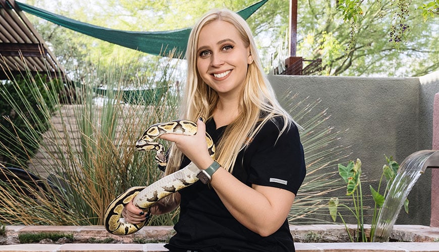 A person with long blonde hair wearing a black shirt is sitting outside, holding a large snake like a seasoned veterinarian. The background includes lush greenery and a stone wall.
