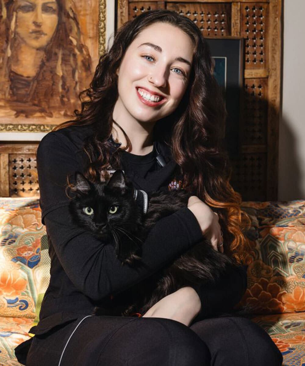 A smiling woman with long brown hair sits on a patterned sofa, holding a black cat in her arms. A framed portrait and wooden panel are visible in the background.